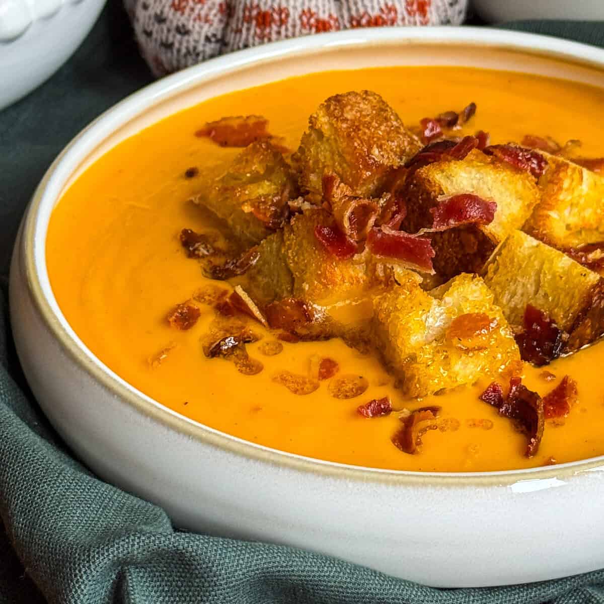 A close-up of creamy butternut squash coconut and chilli soup topped with golden croutons and crispy bacon bits, served in a white bowl on a dark green cloth.