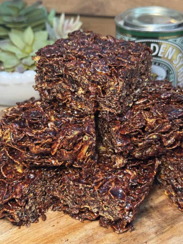 A close-up of chocolate crispy cereal squares stacked on a wooden surface, with a can of golden syrup and a succulent plant blurred in the background.