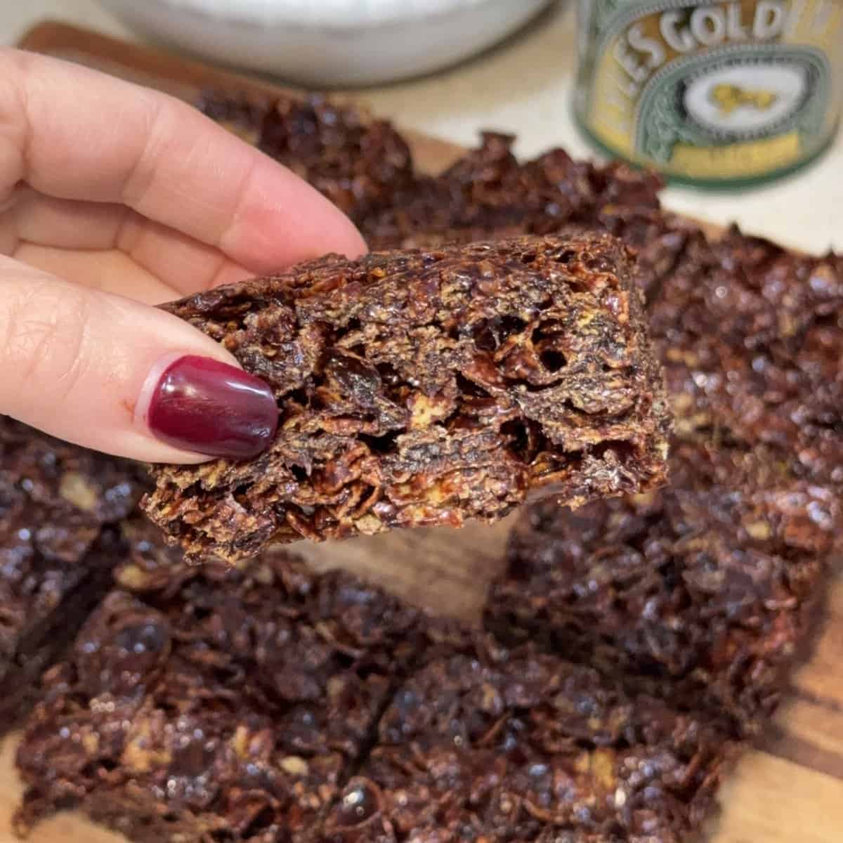A hand with dark red nails holds up a chocolate cracknell square, showing its chewy, textured inside. More flapjack bars are arranged on a wooden board in the background.