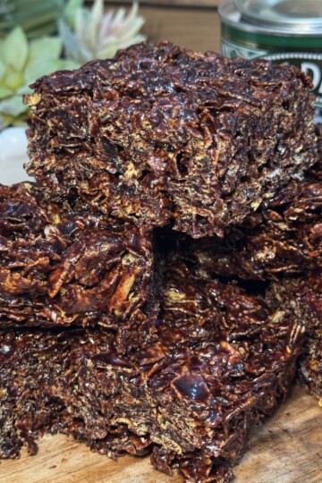 A close-up of chocolate cracknell squares stacked on a wooden surface, with a can of golden syrup and a potted succulent plant in the background.