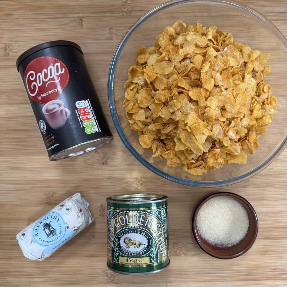 A bowl of cornflakes, a tin of cocoa powder, a can of golden syrup, a small dish of sugar, and a block of butter arranged on a wooden surface.