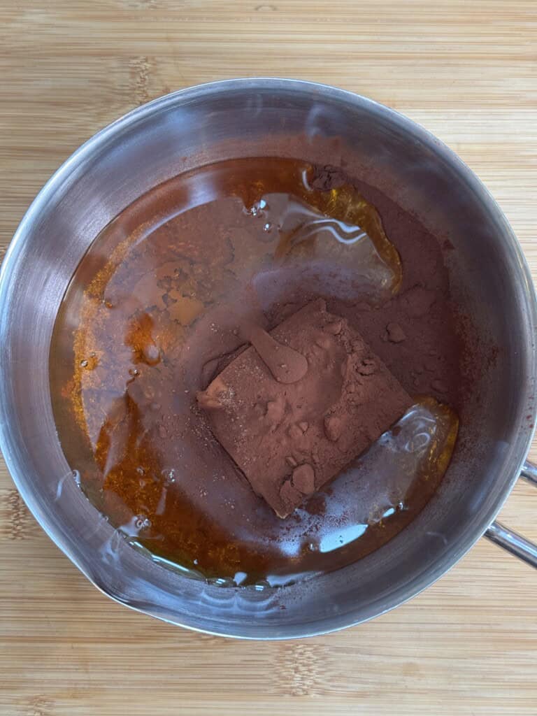 A saucepan on a wooden surface contains melted butter, cocoa powder, and a small spatula, with the ingredients partially mixed.
