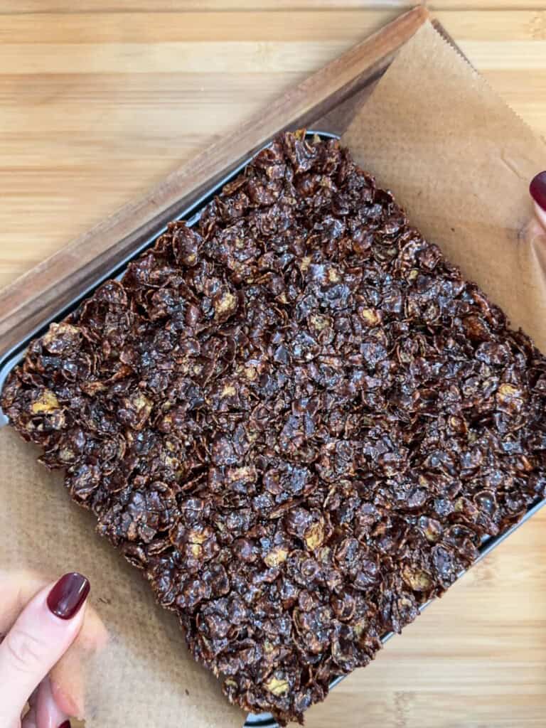 A person lifts a square tray of chocolate cornflake cake from parchment paper on a wooden surface. The cake is textured with visible cornflakes and appears glossy and rich.