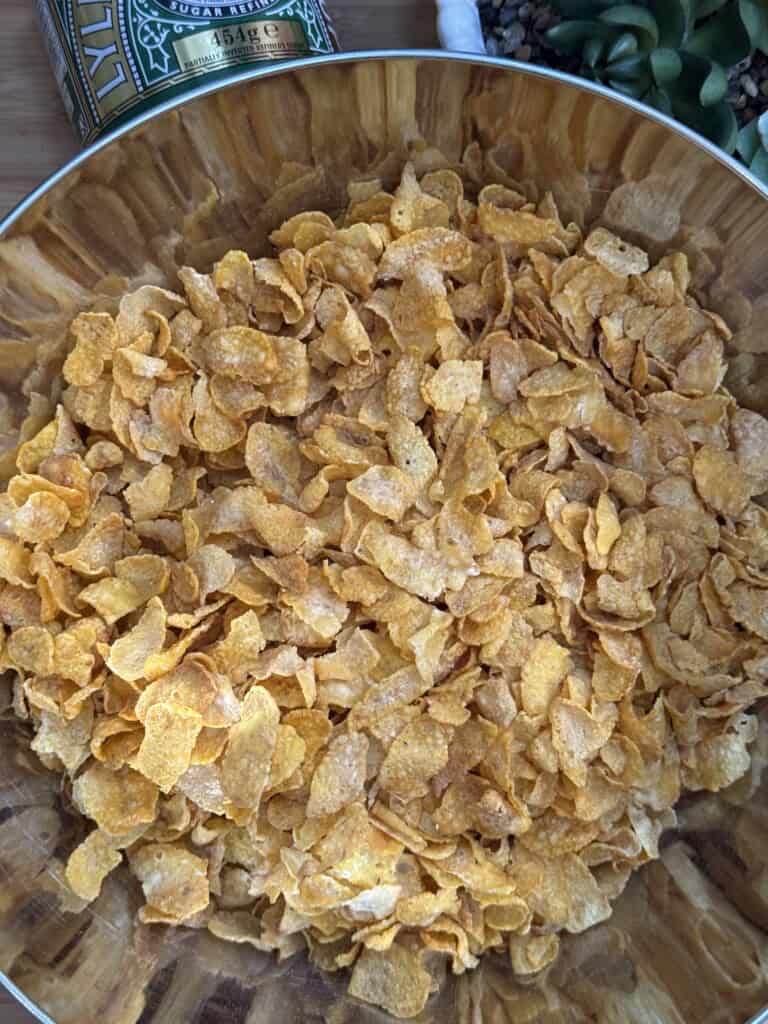 A close-up view of a large wooden bowl filled with corn flakes cereal, with part of a tin container and a succulent plant visible at the edge of the frame.