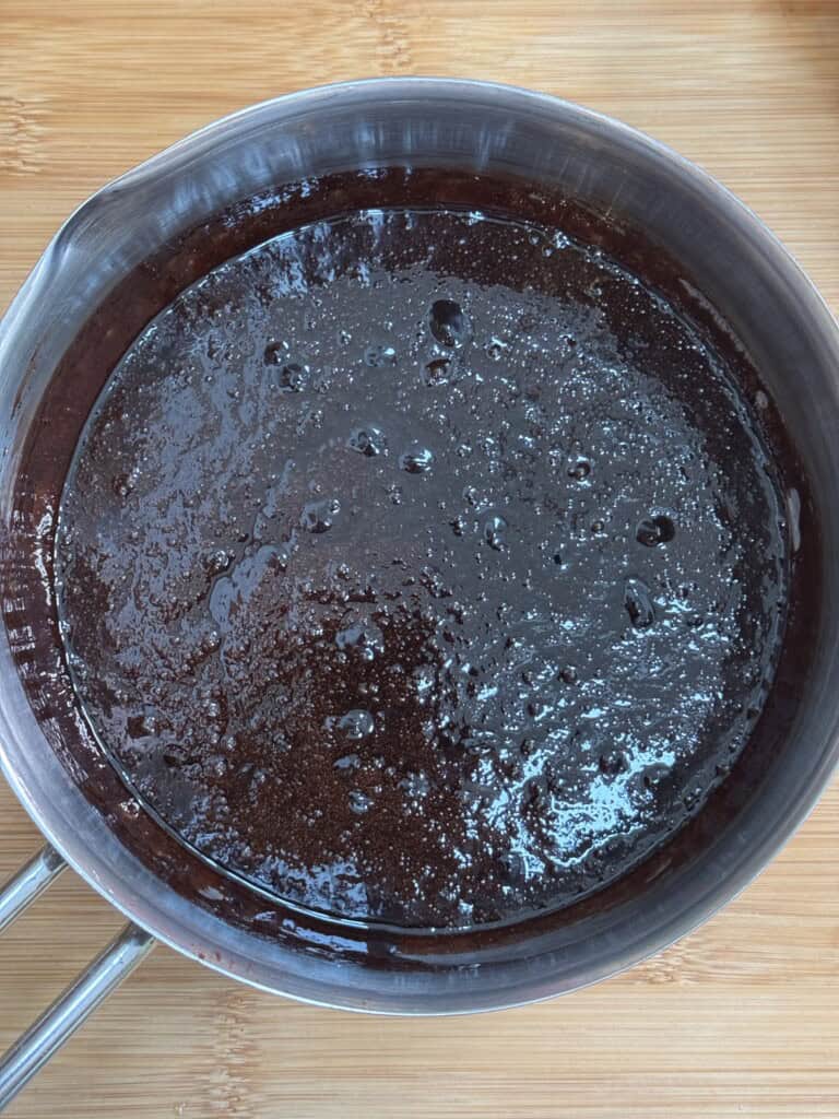 A close-up view of a saucepan filled with glossy, melted chocolate mixture, sitting on a wooden surface. The mixture appears smooth with some bubbles on the surface.