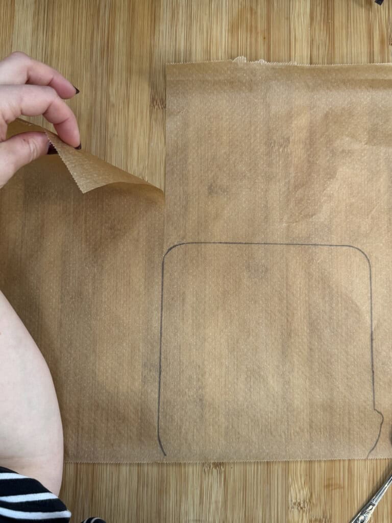 A hand lifts the corner of a piece of brown parchment paper with a square outline drawn on it, placed on a wooden surface.