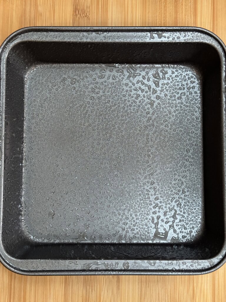 A square metal baking pan lightly coated with oil sits on a wooden surface, ready for use.