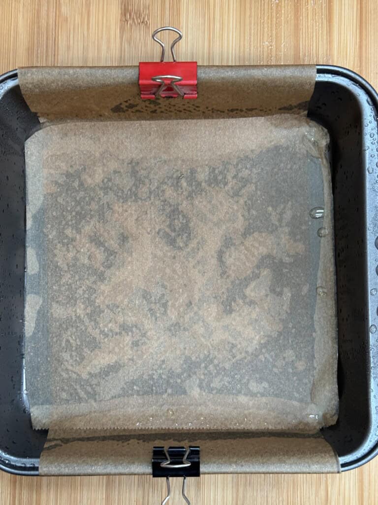 A square baking tin lined with parchment paper, held in place by two binder clips—one red and one black—on opposite sides. The tin sits on a wooden surface.
