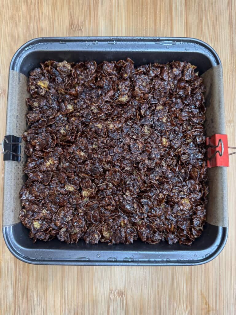 A square baking tin lined with parchment paper, filled with a chocolate cornflake mixture, sits on a wooden surface. The mixture is evenly spread and appears ready to set or bake.