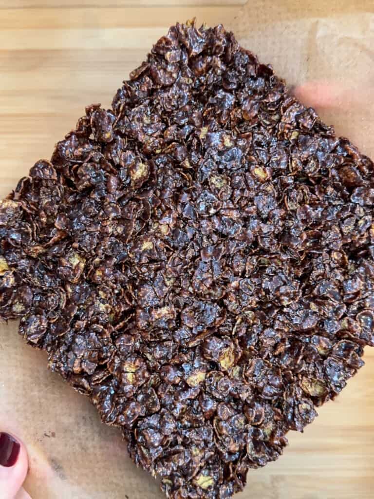 A close-up of a large, square chocolate cracknell on parchment paper, held at the corners by two hands with maroon-painted nails. The surface appears glossy and textured.