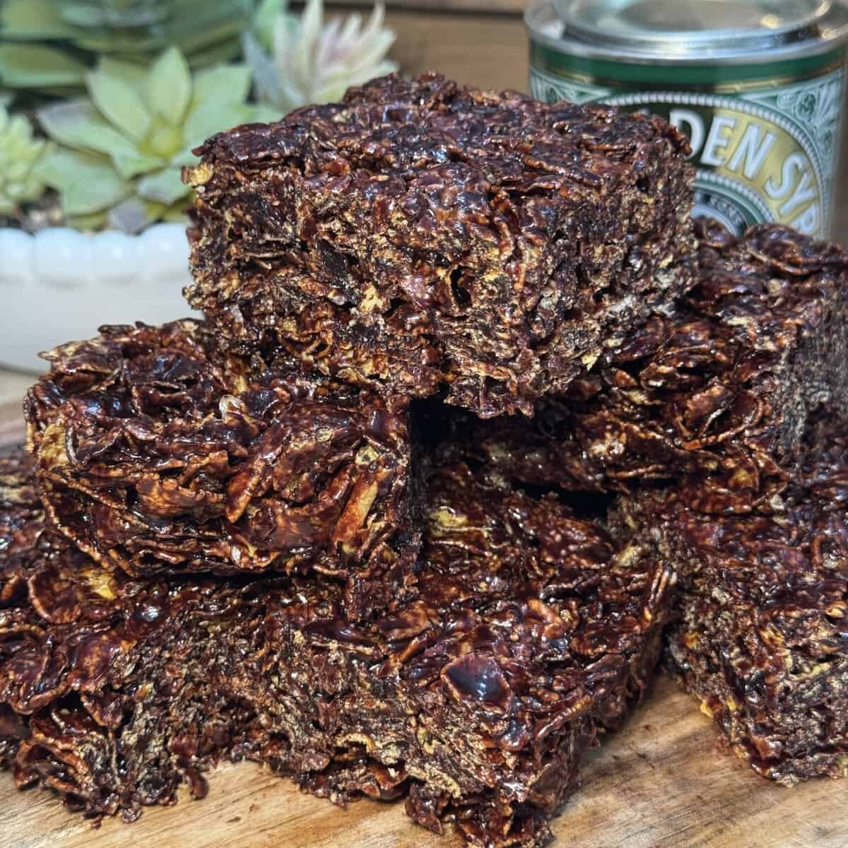 A close-up of chocolate cracknell squares stacked on a wooden surface, with a can of golden syrup and a potted succulent plant in the background.