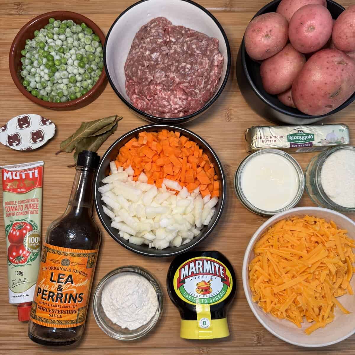 A wooden surface with ingredients for cottage pie: ground meat, diced carrots and onions, peas, potatoes, butter, milk, cheddar cheese, Worcestershire sauce, tomato purée, Marmite, bay leaves, and flour.