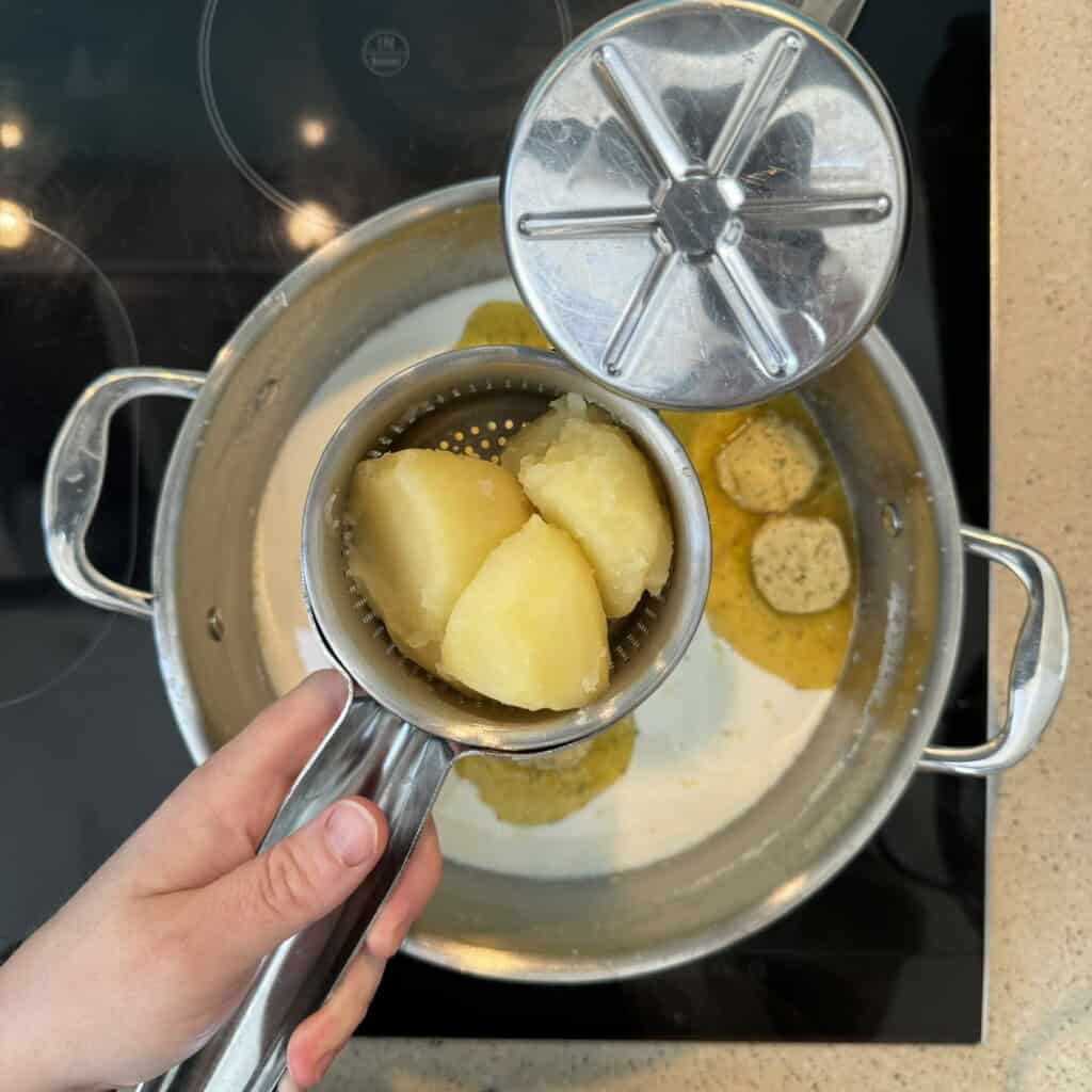 A hand holds a metal potato ricer filled with peeled, boiled potatoes above a saucepan containing melted butter and round slices of herb butter on a stovetop.