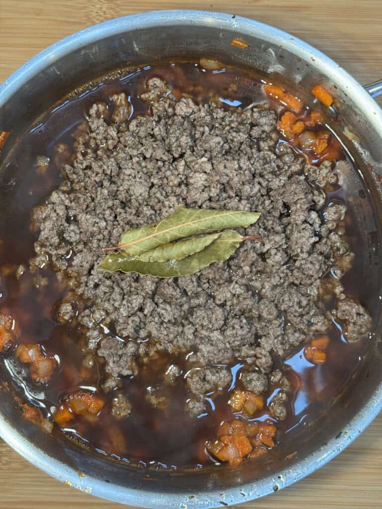 A stainless steel pan filled with ground beef in gravy, diced carrots, and two bay leaves on top, sitting on a wooden surface.