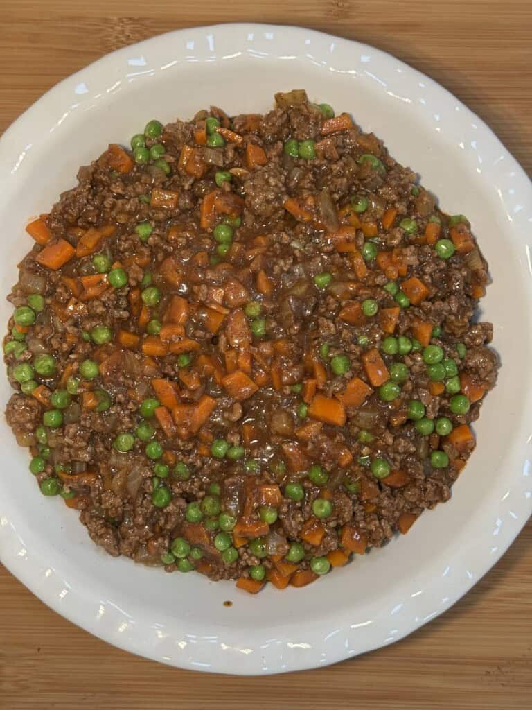 A white plate filled with cottage pie base mix - cooked ground beef mixed with peas, diced carrots, and onions in a brown sauce, placed on a wooden surface.