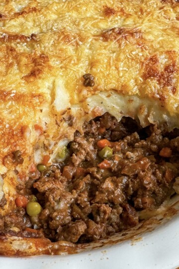 A close-up of a homemade cottage pie in a white dish, showing a golden, cheesy mashed potato topping with a section cut out, revealing ground meat, peas, carrots, and gravy inside.