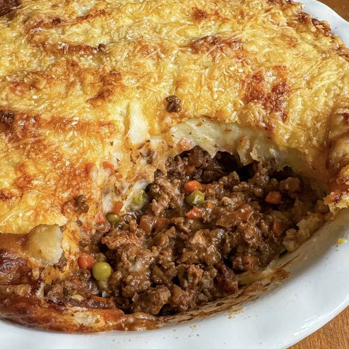 A close-up of a homemade cottage pie with cheese in a white dish, showing a golden, cheesy mashed potato topping with a section cut out, revealing ground meat, peas, carrots, and gravy inside.