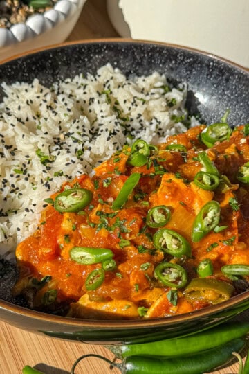 A black bowl filled with white rice topped with black sesame seeds and a vibrant orange curry garnished with sliced green chilies and herbs, placed on a wooden surface with whole green chilies nearby.