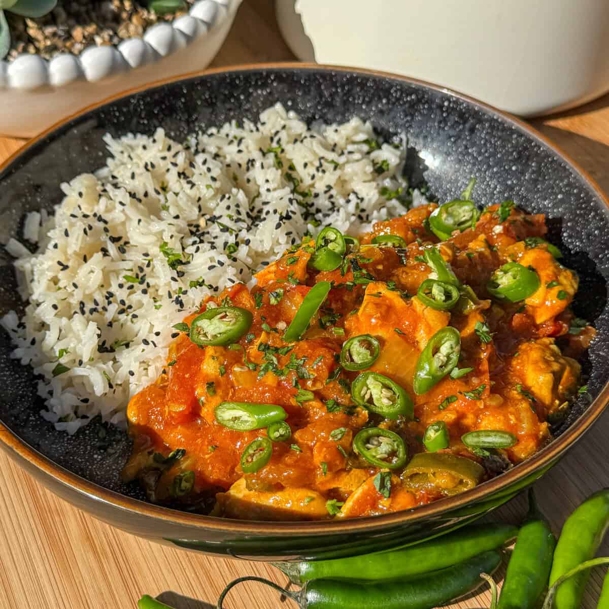 A black bowl filled with white rice topped with black sesame seeds and a vibrant South Indian garlic chilli chicken curry garnished with sliced green chilies and herbs, placed on a wooden surface with whole green chilies nearby.
