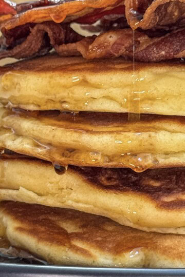 A stack of fluffy pancakes topped with crispy bacon, drizzled with maple syrup, sits on a black plate.