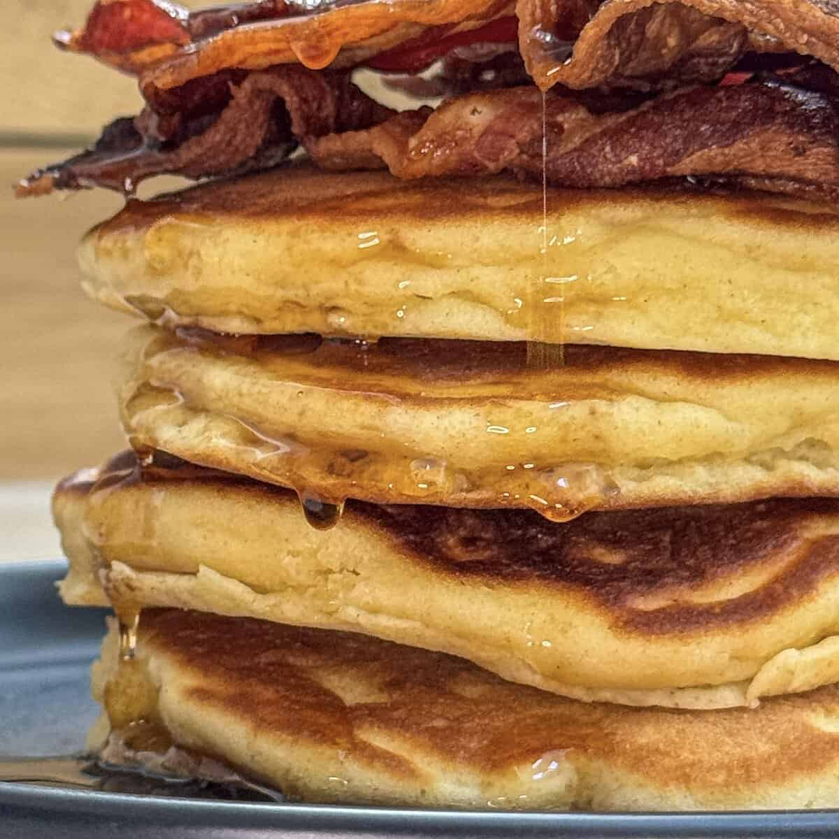 A stack of fluffy pancakes topped with crispy bacon, drizzled with maple syrup, sits on a black plate.