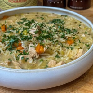 A white bowl filled with chicken noodle soup, featuring chunks of chicken, carrots, celery, noodles, and garnished with chopped parsley. The background shows condiment containers slightly out of focus.