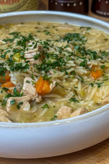 A white bowl filled with chicken noodle soup, featuring chunks of chicken, carrots, celery, noodles, and garnished with chopped parsley. The background shows condiment containers slightly out of focus.