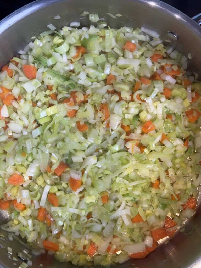Chopped onions, carrots, and celery sautéing in a large stainless steel pan, starting to soften and mix together.