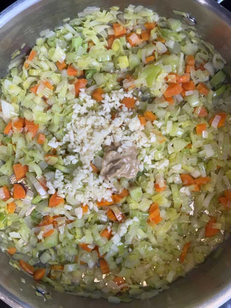Chopped onions, carrots, and leeks are sautéing in a pan. A small mound of minced garlic and a dollop of light brown paste, likely miso or peanut butter, are in the center.