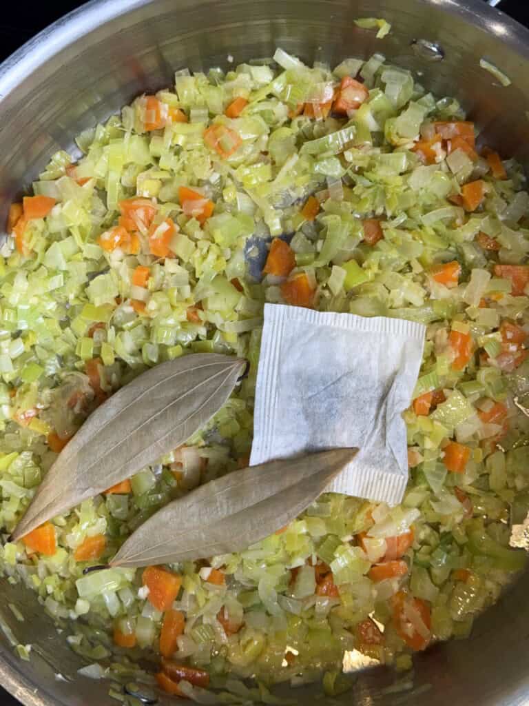Chopped onions, carrots, and celery cooking in a pan with two bay leaves and a spice sachet on top.