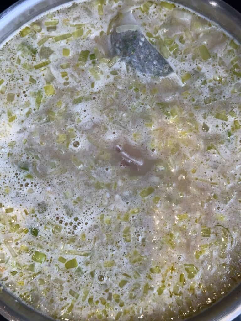 A close-up of a pot filled with a simmering creamy soup, likely containing chopped leeks, onions, and herbs, with bubbles forming on the surface.