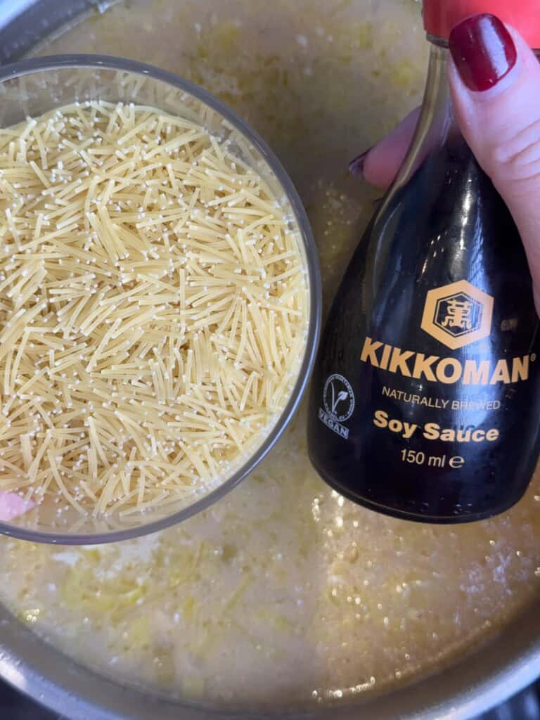A hand holds a bowl of uncooked short pasta over a pot of simmering soup, alongside a bottle of Kikkoman soy sauce. The soup appears creamy with visible pieces of vegetables.