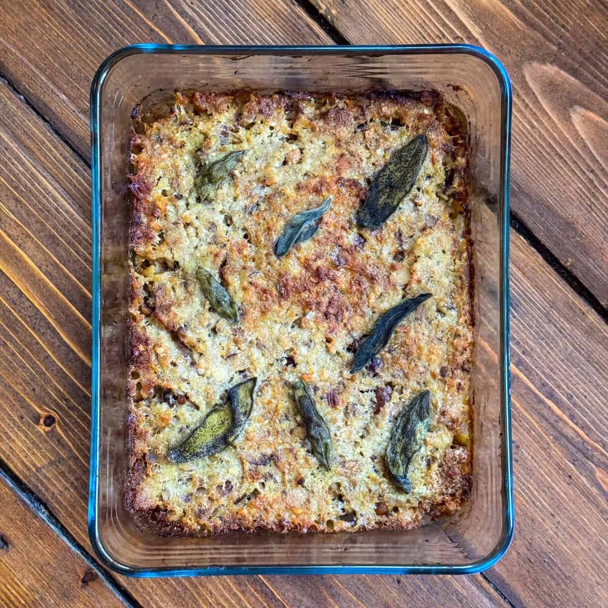 A glass baking dish filled with sausage meat stuffing with paxo topped with whole sage leaves, sitting on a wooden surface.