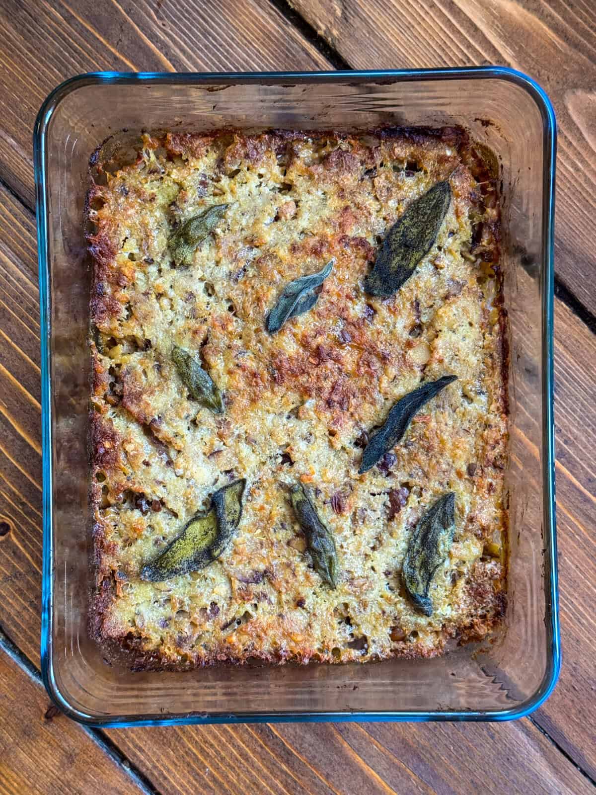 A glass baking dish filled with golden brown sausage meat stuffing with paxo, topped with several whole sage leaves, sits on a wooden surface.