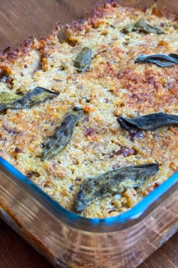 Baked sausage meat stuffing with paxo with a golden-brown, crispy top, dotted with browned sage leaves, sits on a wooden surface. The edges appear slightly caramelized.