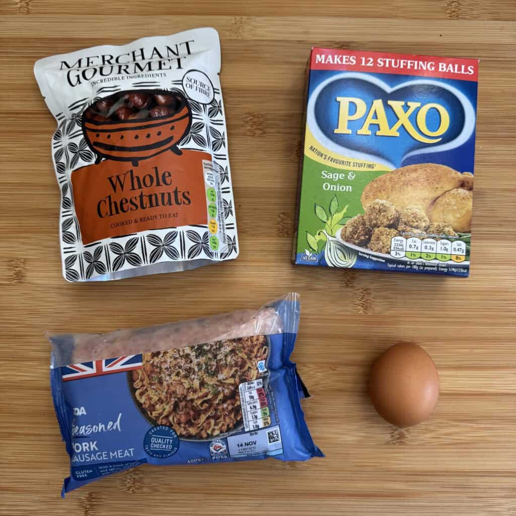 Four items on a wooden surface: a bag of whole chestnuts, a box of Paxo sage and onion stuffing mix, a pack of seasoned pork sausage meat, and a single brown egg.