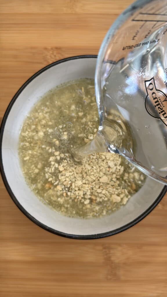 A glass measuring cup pours water into a white bowl filled with dry instant oatmeal, set on a wooden surface.