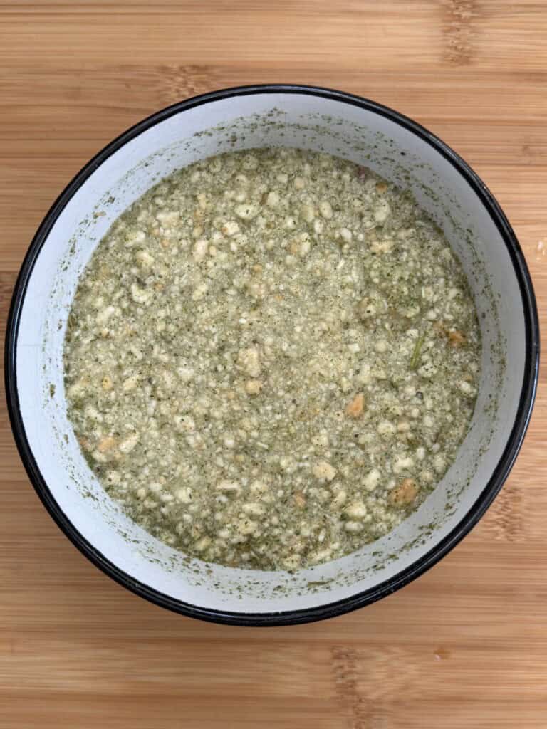 A white bowl with a black rim filled with a chunky, greenish mixture sits on a wooden surface. The mixture appears to contain herbs and small bits of bread or crackers.