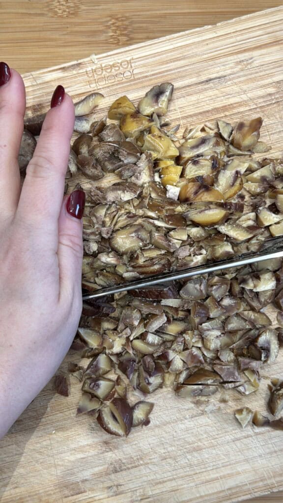 A hand with dark red nail polish holds chopped chestnuts on a wooden cutting board while a knife slices through them.