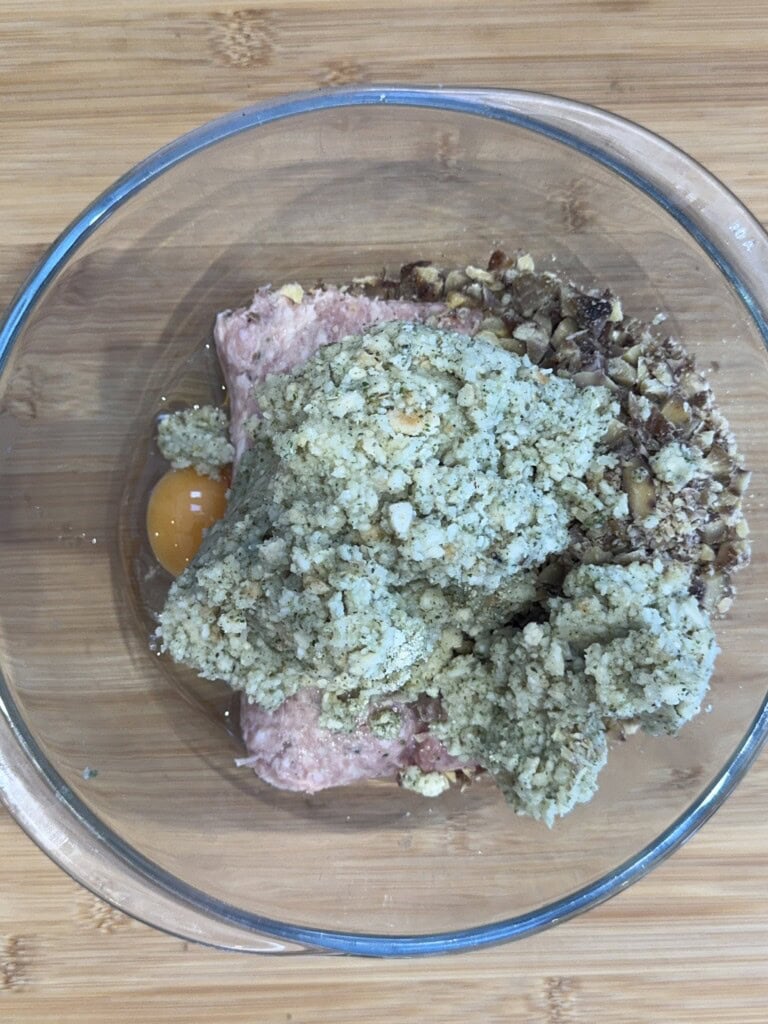 A glass bowl containing ground meat, a raw egg, chopped nuts, and a pile of breadcrumb mixture, all sitting on a wooden surface.
