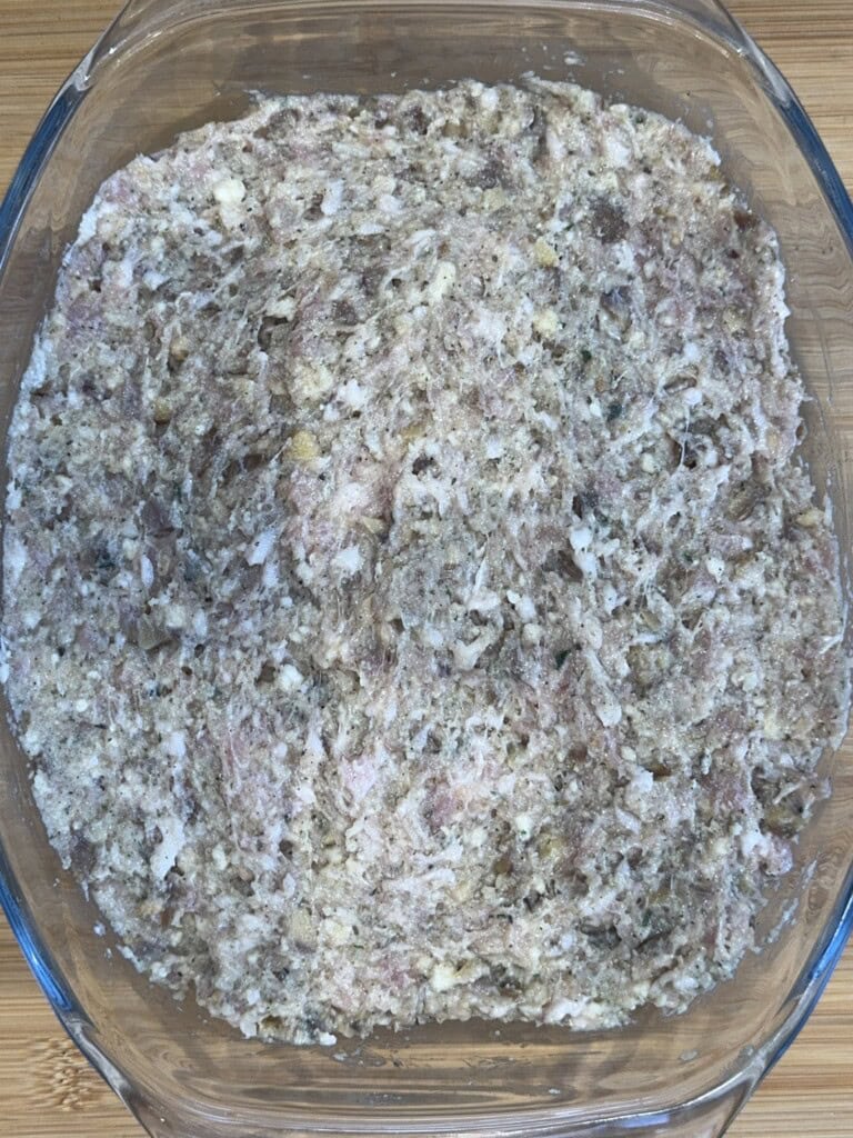 A glass dish filled with a raw, mixed meatloaf mixture containing ground meat, breadcrumbs, and seasonings, ready to be shaped or cooked. The mixture has a rough, uneven texture.