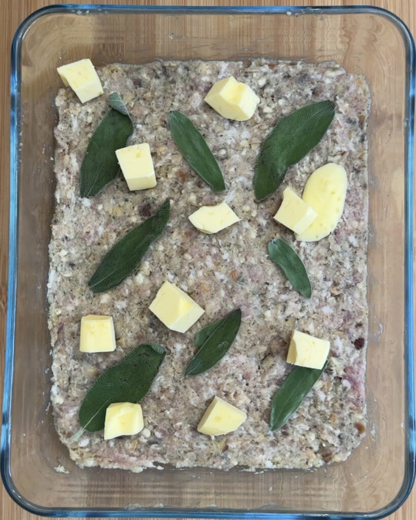 A glass oven dish containing an uncooked meat mixture topped with fresh sage leaves and small cubes of butter, set on a wooden surface.