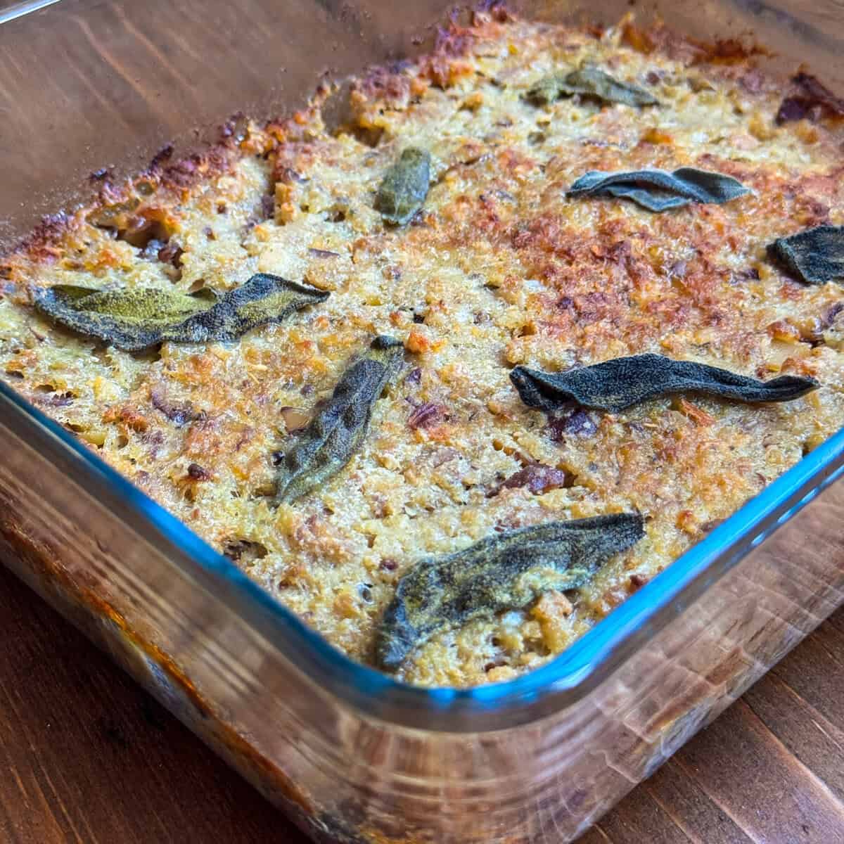 Baked sausage meat stuffing with paxo with a golden-brown, crispy top, dotted with browned sage leaves, sits on a wooden surface. The edges appear slightly caramelized.