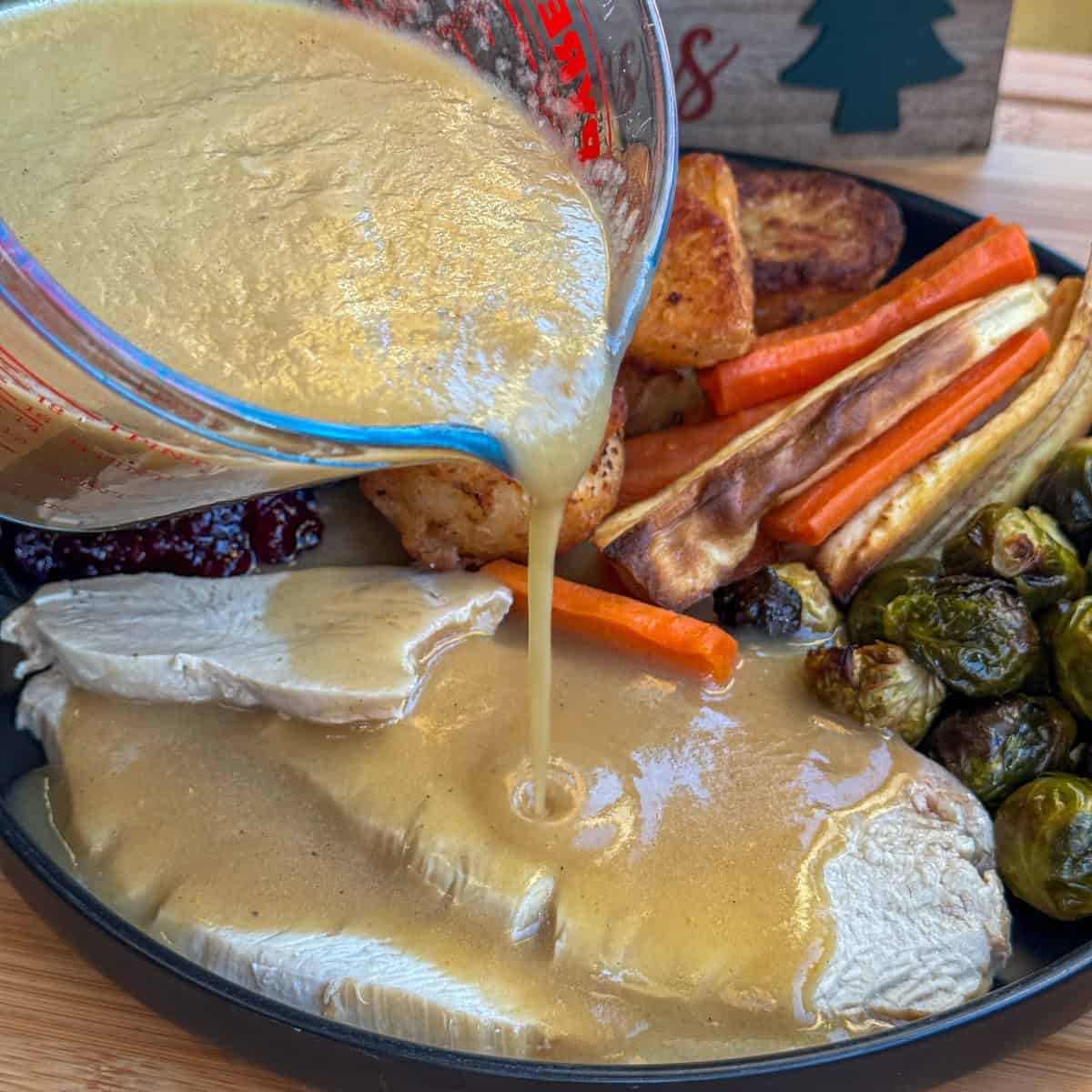 A hand pours christmas gravy from a measuring jug over slices of roasted turkey, surrounded by roasted potatoes, carrots, parsnips, Brussels sprouts, and cranberry sauce on a plate.
