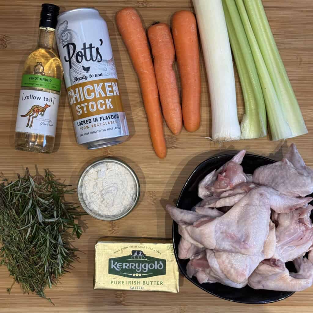 A wooden surface displays fresh rosemary, a small wine bottle, chicken stock can, three carrots, two leeks, two celery stalks, flour in a dish, butter, and raw chicken wings in a black bowl.