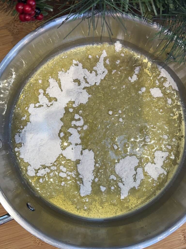 A metal pan containing melted butter with flour sprinkled on top, ready to be mixed. Holiday greenery and red berries decorate the wooden surface around the pan.