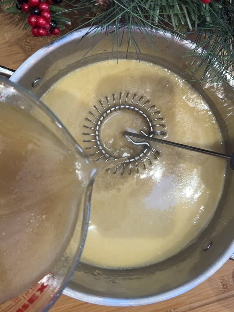 A pot of light yellow mixture is being whisked while a liquid is poured in from a measuring cup; holiday greenery and red berries are visible at the top of the image.