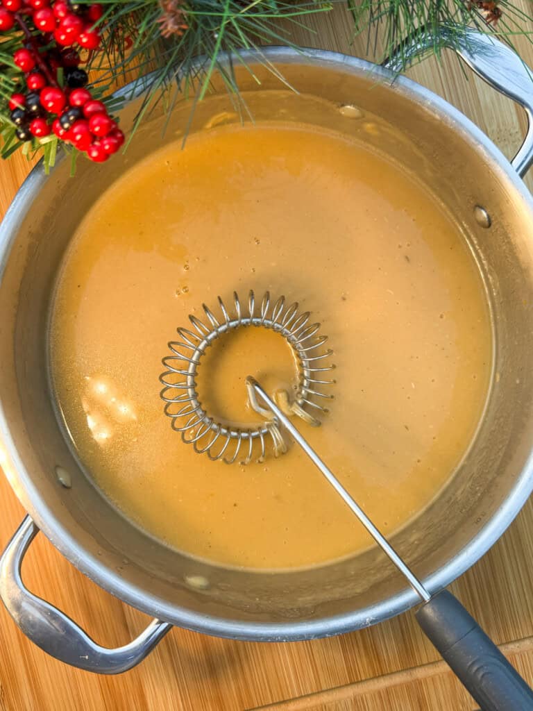 A pot of smooth brown gravy being whisked with a metal spiral whisk, set on a wooden surface with holiday greenery and red berries in the corner.