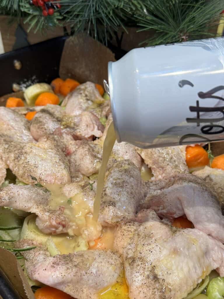 Raw chicken wings and vegetables are in a roasting pan, seasoned with pepper. A hand pours beer from a can over the ingredients. Pine branches and red berries are visible in the background.