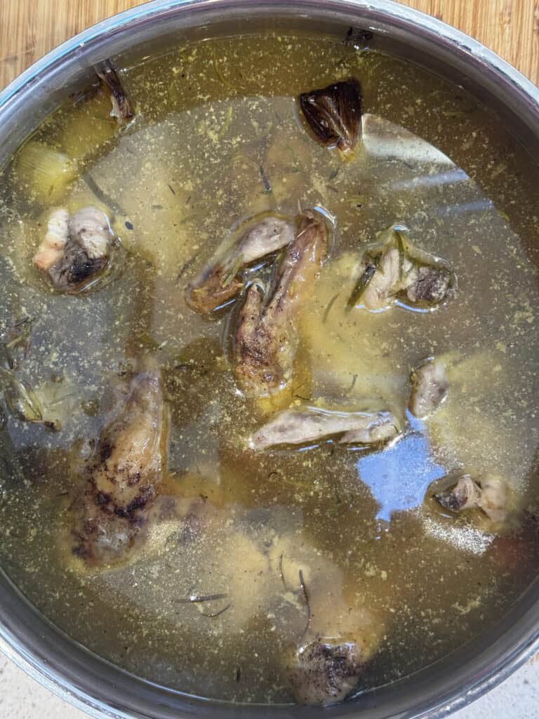 A pot of homemade chicken broth with chicken pieces, herbs, and vegetables floating in clear golden broth on a wooden surface.